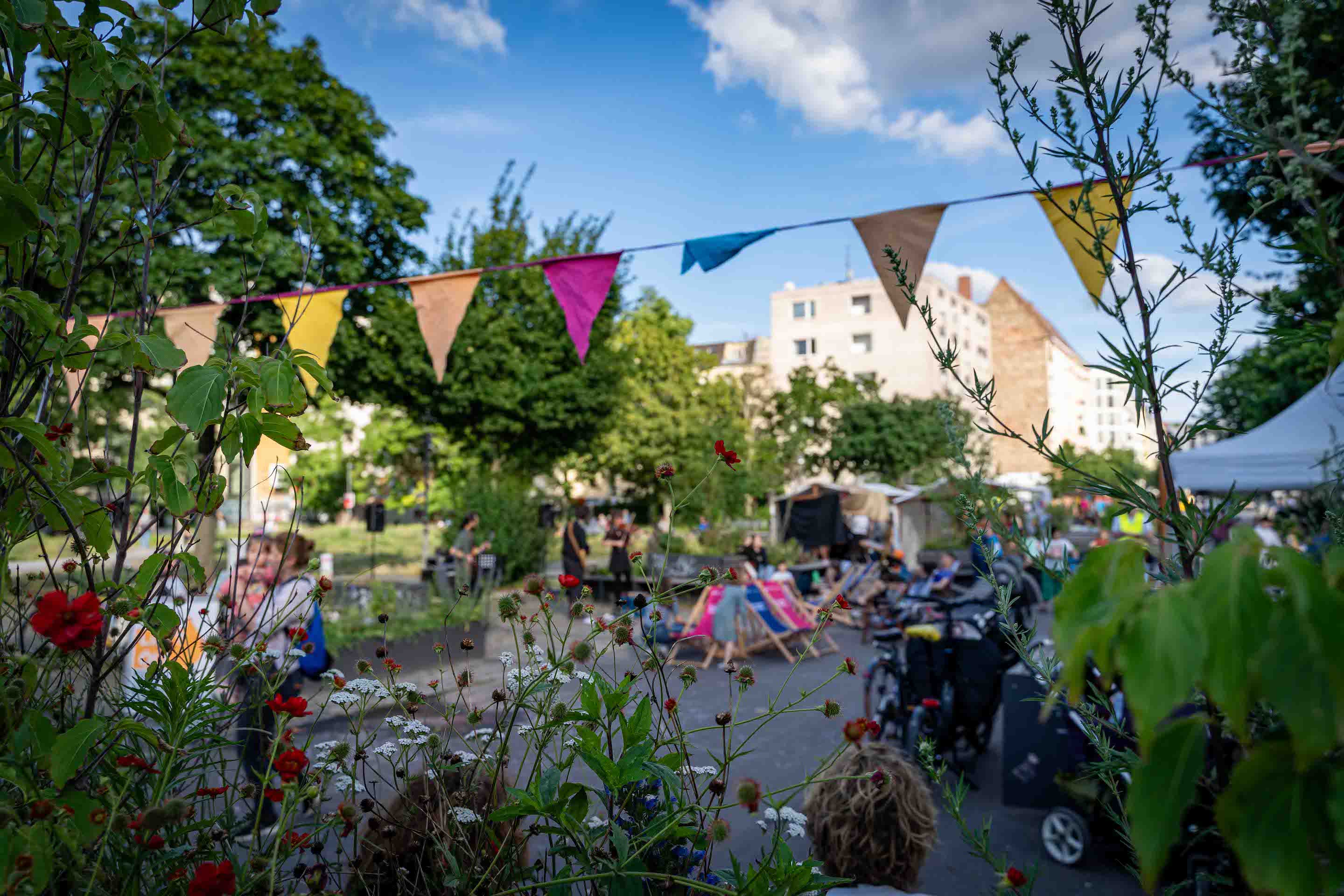 Nachhaltige Stadtentwicklung für starke Nachbarschaften: Impressionen vom Tag des guten Lebens 2025. Man sieht bunte Wimpel, Pflanzen und Menschen vor Liegestühlen und Lastenrädern.